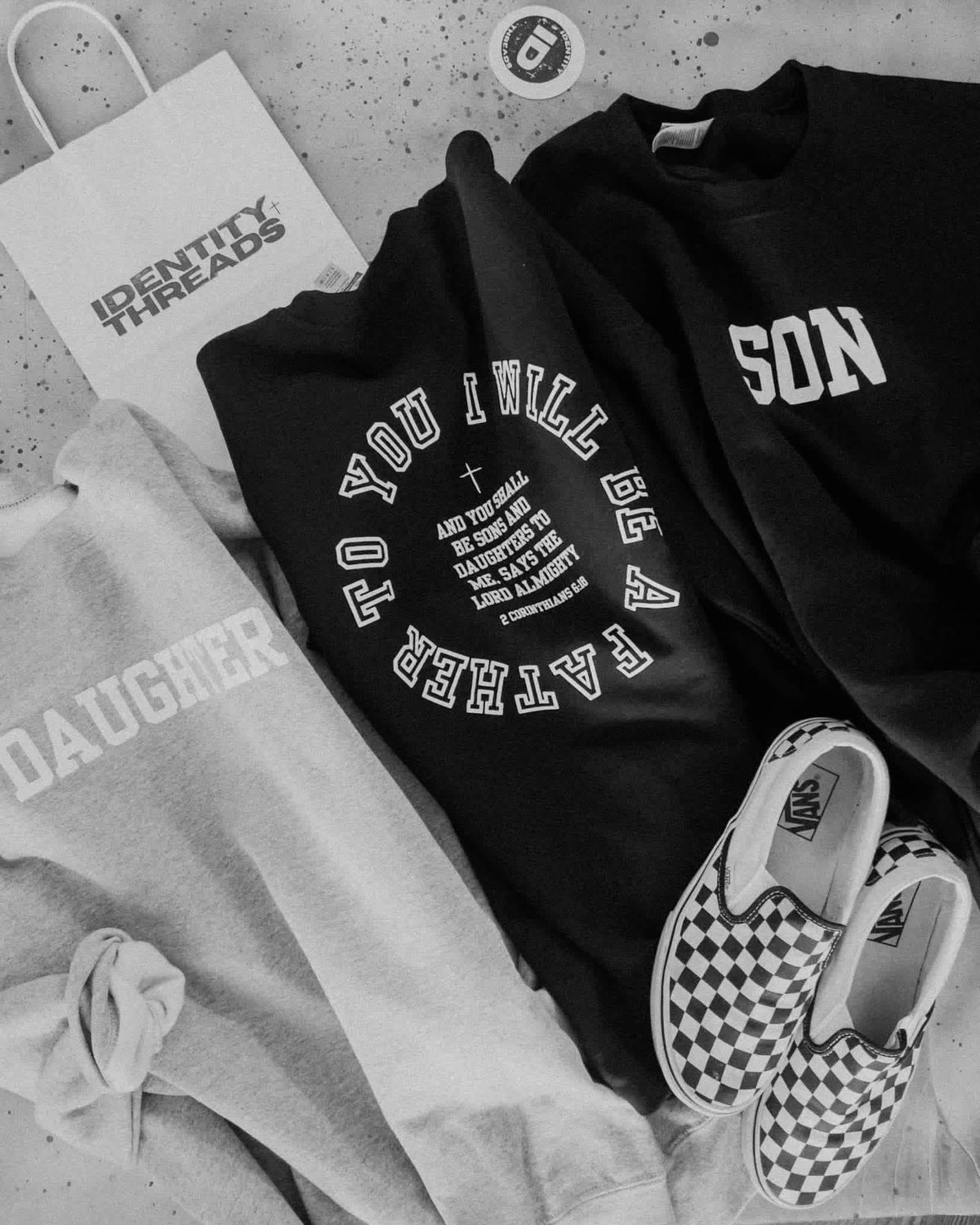 A styled flat-lay of black and gray graphic sweatshirts with white text, a white paper shopping bag reading 'IDENTITY THREADS,' a small round sticker, and black-and-white checkerboard slip-on sneakers on a speckled surface.