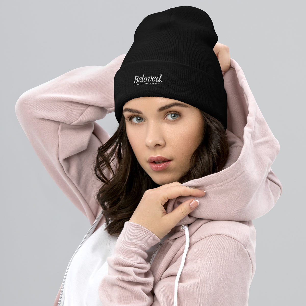 Woman wearing a black Christian beanie with a logo saying "beloved" and a pink hoodie on a gray background